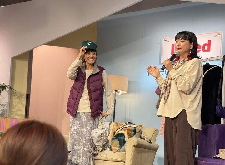 ステージでマイクを持ち話す2人の女性。左はダウンベストとキャップ姿。