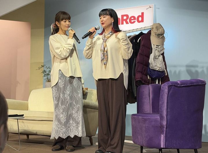 ステージでマイクを持ち話す2人の女性。左はレーススカート、右はスカーフ着用。
