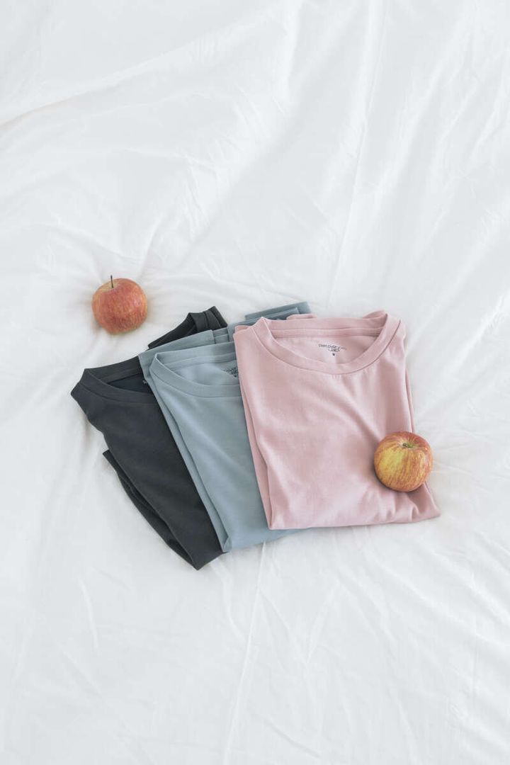 白いシーツに畳まれたピンク、水色、グレー、黒のTシャツとリンゴ2つ
