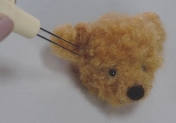 羊毛フェルトで作られたクマの顔の耳部分をニードルで刺している手元