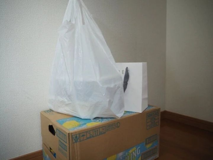 玄関の床に置かれた段ボール箱の上に、白いレジ袋と白い紙袋が積まれている
