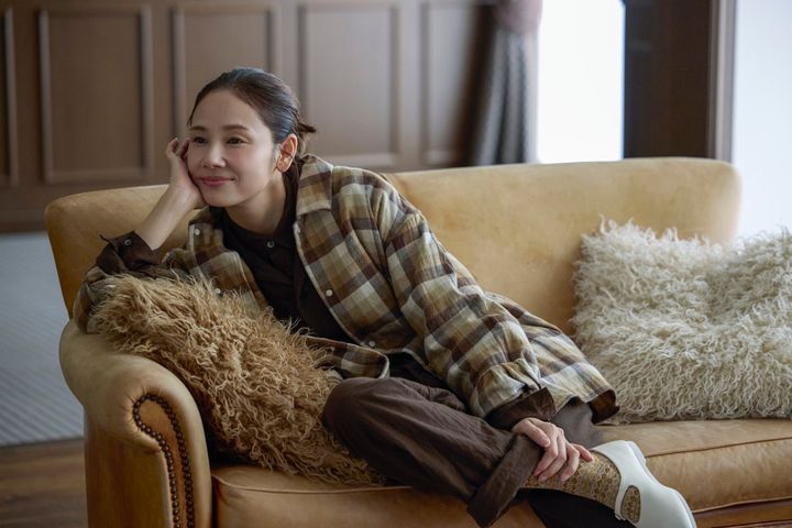 チェック柄の上着と茶系のパンツを合わせ、ソファに座る吉田羊さん