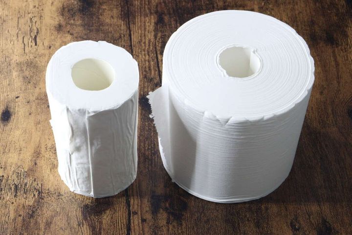 木目のテーブルに、左に圧縮トイレットペーパー、右に通常の5倍巻きトイレットペーパーが並べられている。