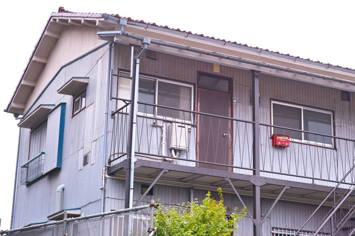 古い二階建てアパートの外観。二階の玄関ドアのそばに赤い郵便受けが設置されている。