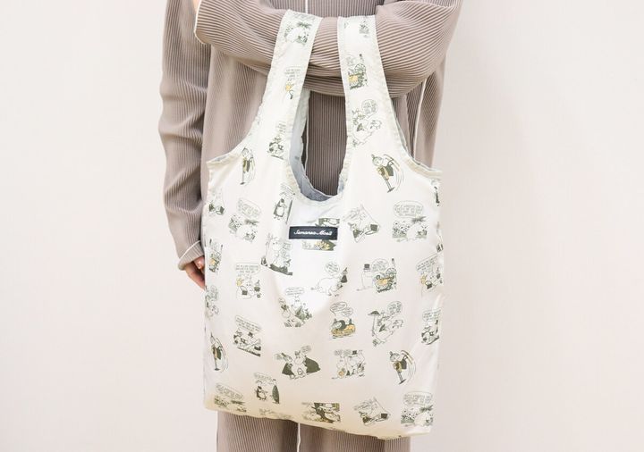 グレーの服を着た人物が、ムーミンのコミック柄エコバッグを肩にかけている全身ショット。バッグは広げられた状態。