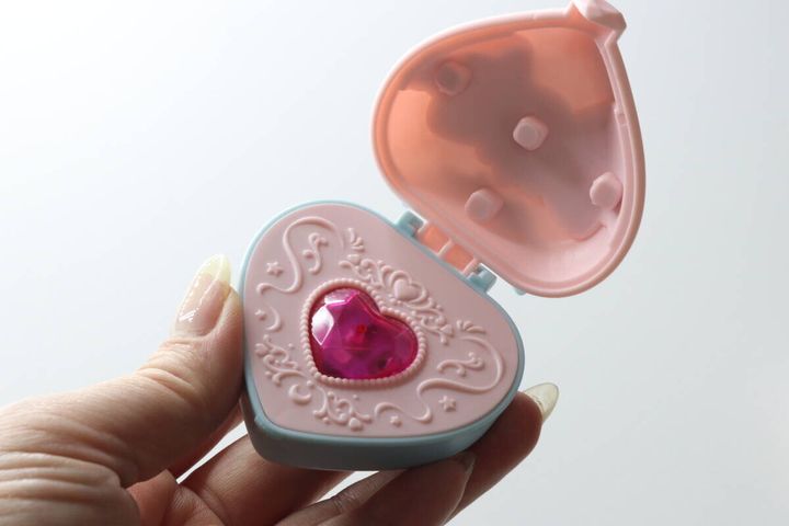 女性の手が持っている、蓋が開いた状態のハート型マジカルフラッシュコンパクト。内側はピンク色で中央に赤いストーンがある
