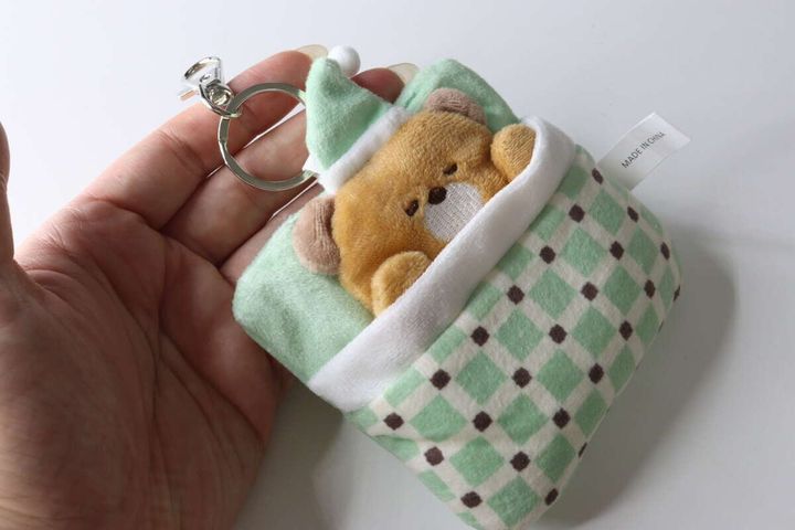 女性の手のひらに、緑の布団に入った茶色いくまのキーホルダーが乗っている