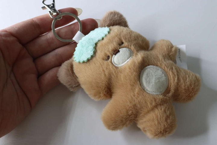女性の手のひらに、アイマスクをした茶色いくまのキーホルダーが乗っている