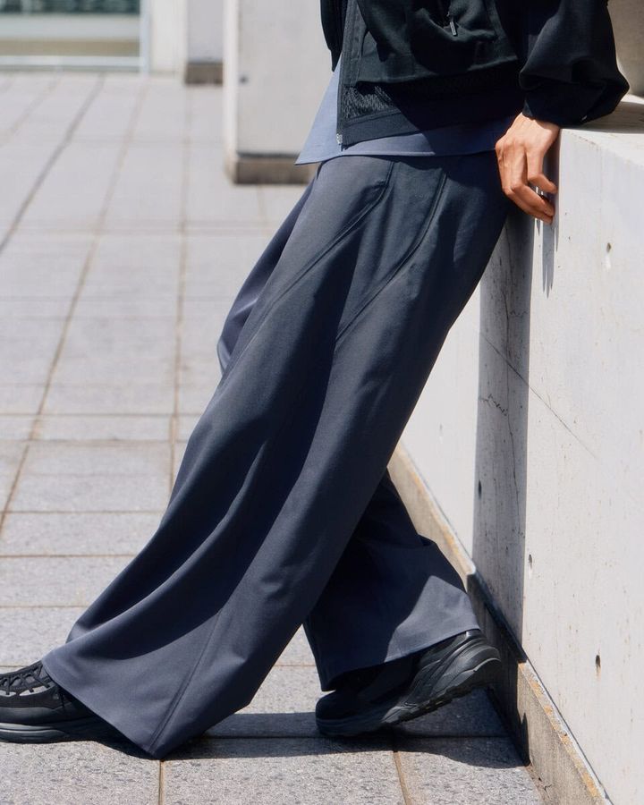 ノータックのすっきりとした腰まわりから、裾までまっすぐに落ちるワイドストレートパンツ。身体のラインを拾いにくい適度なゆとりと、ウールのしなやかなドレープが端正なフォルムをつくっている。