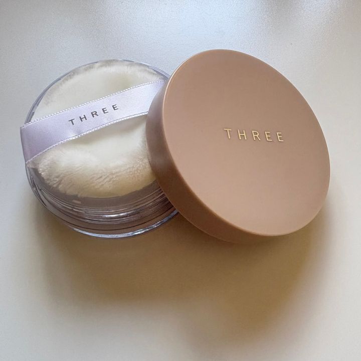 「THREE」のブランドロゴがゴールドで刻印されたベージュゴールドの蓋が、クリアな丸型容器の横に立てかけられた、「THREE クラウドヴェール ルースパウダー X02」の限定パッケージ。容器の中には、「THREE」のロゴ入り白いリボンが付いたふわふわとした白いフェイスパフが収められています。