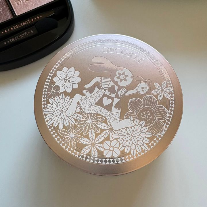 コスメデコルテ限定フェイスパウダーの天面デザイン。ピンクゴールドの蓋に、不思議の国のアリスをイメージした時計を持つうさぎや満開の花々、レース風の繊細なイラストが白とシルバーの線画で描かれています。
