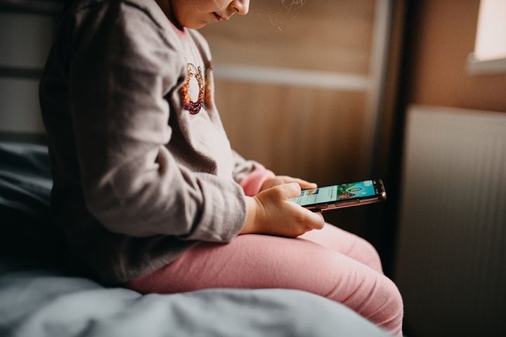 ベッドに座り、スマートフォンに見入る少女です。画面を延々とスクロールするうちに頭が腐っていくように感じる様子が、ブレインロットという言葉の意味です。