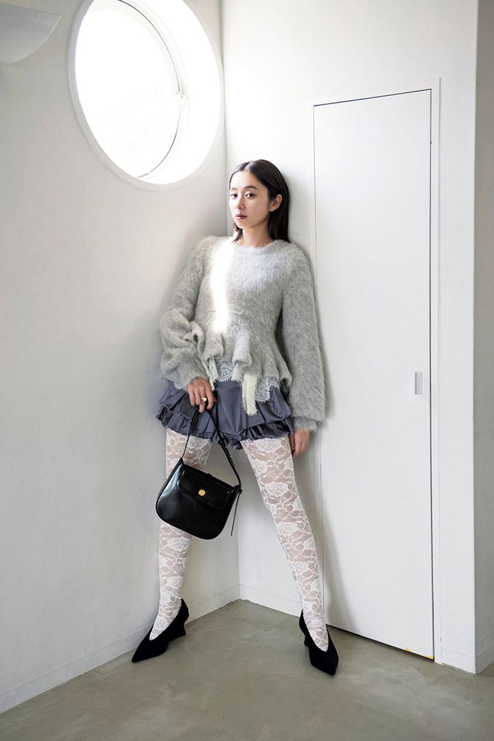 ボリュームのあるグレーのペプラム風ニットに、フリルのショートパンツ、総レースの白いタイツ、黒のパンプスとハンドバッグを合わせた、室内で壁に寄り添う堀田真由。