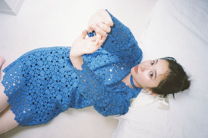 青い総レースのミニドレスに、同色の首元のチョーカーと白いヘアリボンを合わせ、白いベッドの上で横たわってこちらを見つめる堀田真由。