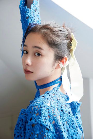青いレースドレスを着用し、白いリボンのヘアアクセサリーと花のイヤーカフをつけ、アンニュイな表情で振り返る堀田真由のアップ。