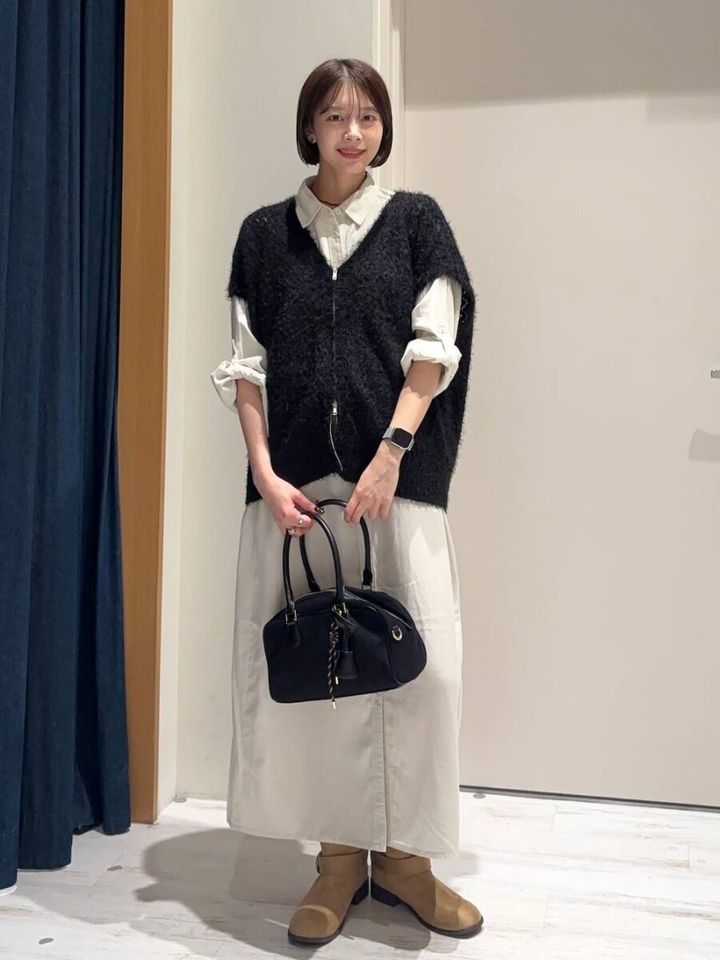 白のシャツワンピースに黒のシャギーベストを重ね黒いバッグを持つショートヘアの女性
