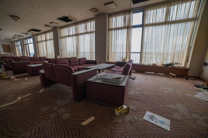 大きな窓にカーテンがかかった、廃旅館のラウンジ。紫色のソファが並び、床には紙くずなどが散乱している。