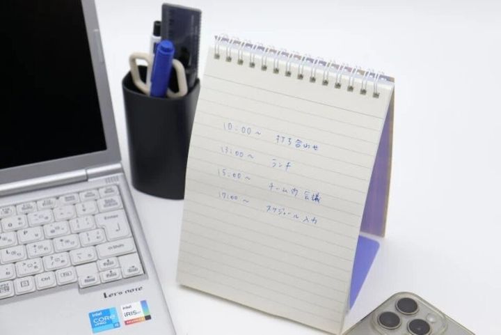 PCの横にスケジュールが書き込まれたスタンドリングノートが立てられている様子