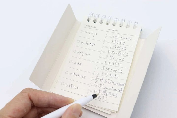 開いた単語帳にペンを持ちながら英単語と意味を書き込んでいる手のアップ写真